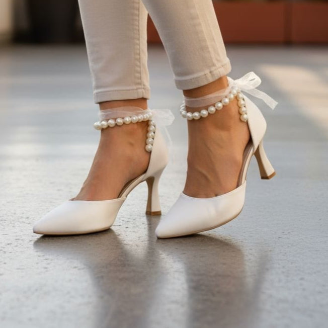 White high-heeled shoes with pearl ankle straps worn by a person on a rooftop.