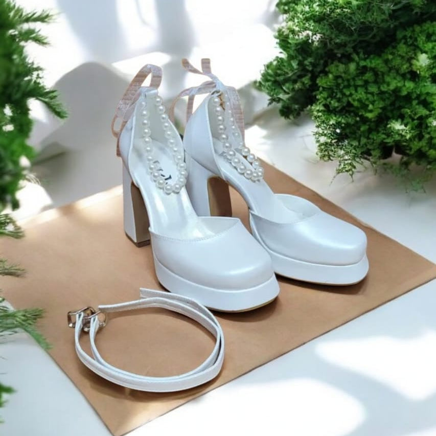 White high-heeled shoes with decorative straps on a brown paper background with green plants.