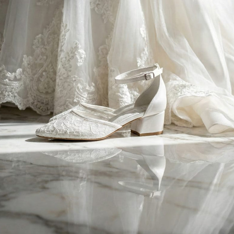 White lace bridal shoe with a low block heel and ankle strap featuring a pointed toe, posed next to a wedding veil.